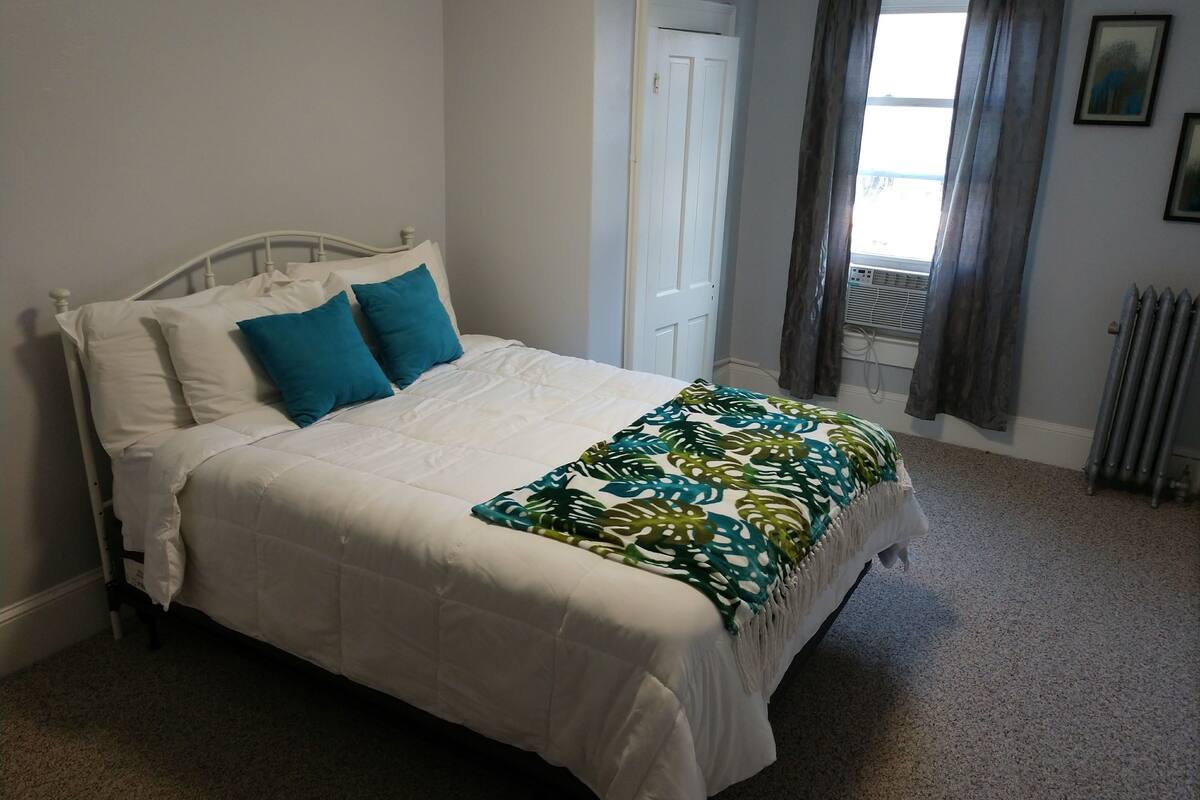 A neatly made bed with white bedding and blue pillows, a radiator, and a window with curtains.