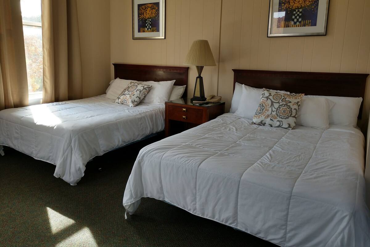 A hotel room with two single beds, a nightstand with a lamp, and two framed artworks on the wall.