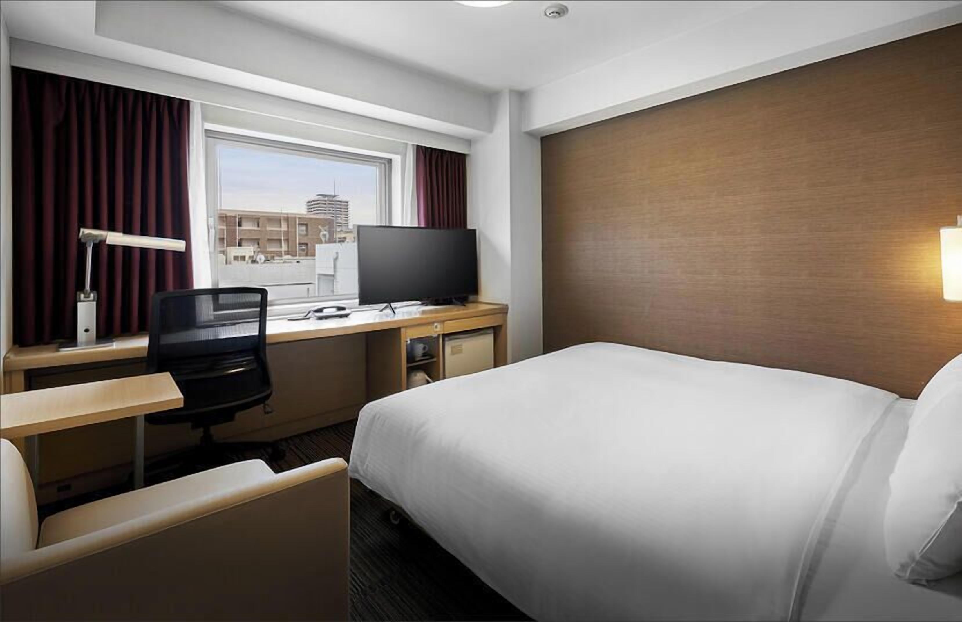 A hotel room with a bed, desk, chair, TV, and a window with curtains.