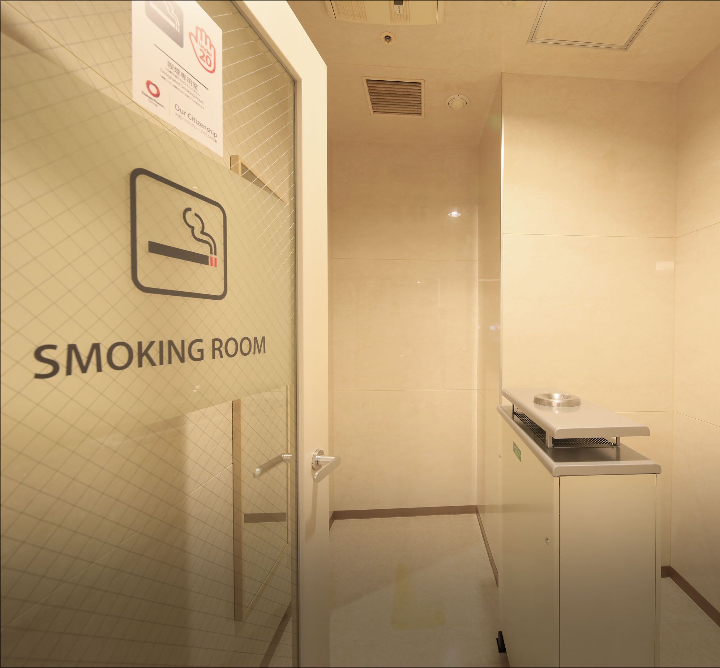 A smoking room with a glass door, a sign indicating it is a smoking area, and a ventilation unit on the wall.