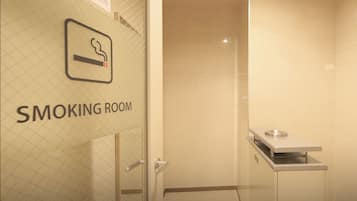 A smoking room with a glass door, a sign indicating it is a smoking area, and a ventilation unit on the wall.