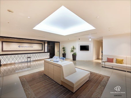 A modern hotel lobby with a reception desk, a seating area, a large TV, and a skylight.