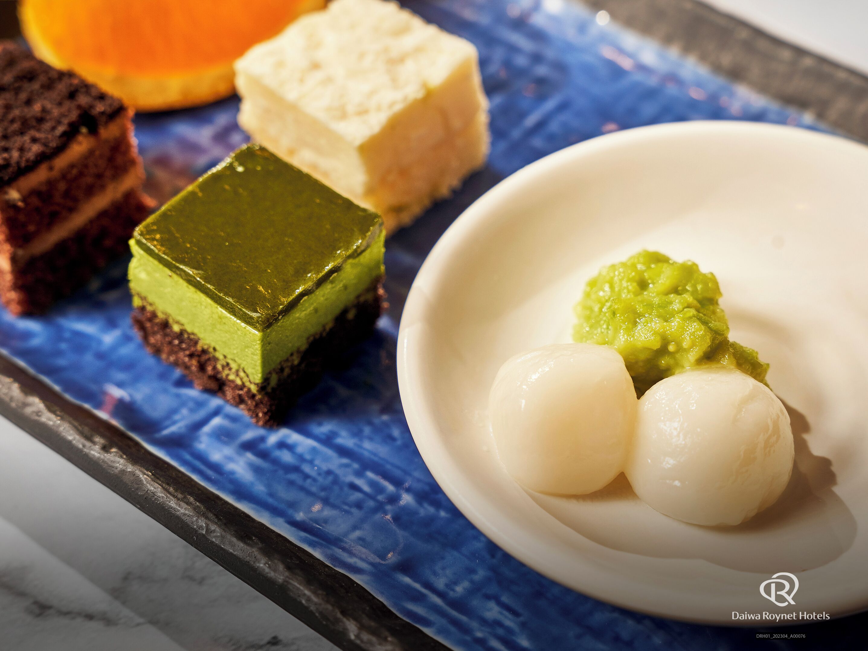 A dessert plate with various sweets, including a green square and two white round items, on a blue cloth.