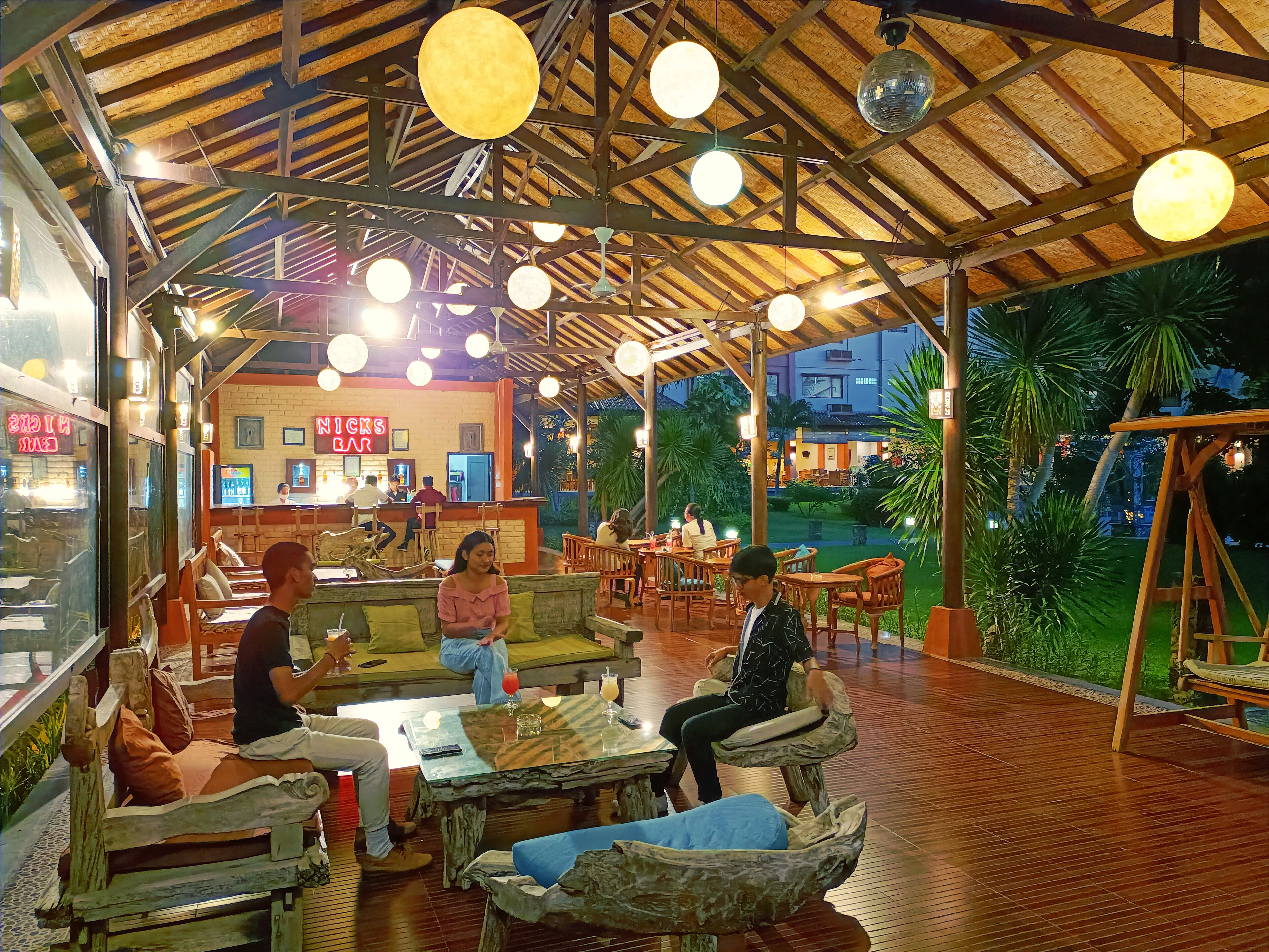 Un restaurante al aire libre con mesas y bancos de madera, techo de paja y luces esféricas colgantes.