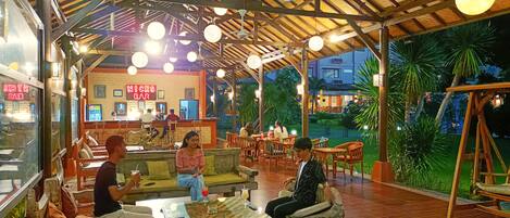 Un restaurante al aire libre con mesas y bancos de madera, techo de paja y luces esféricas colgantes.