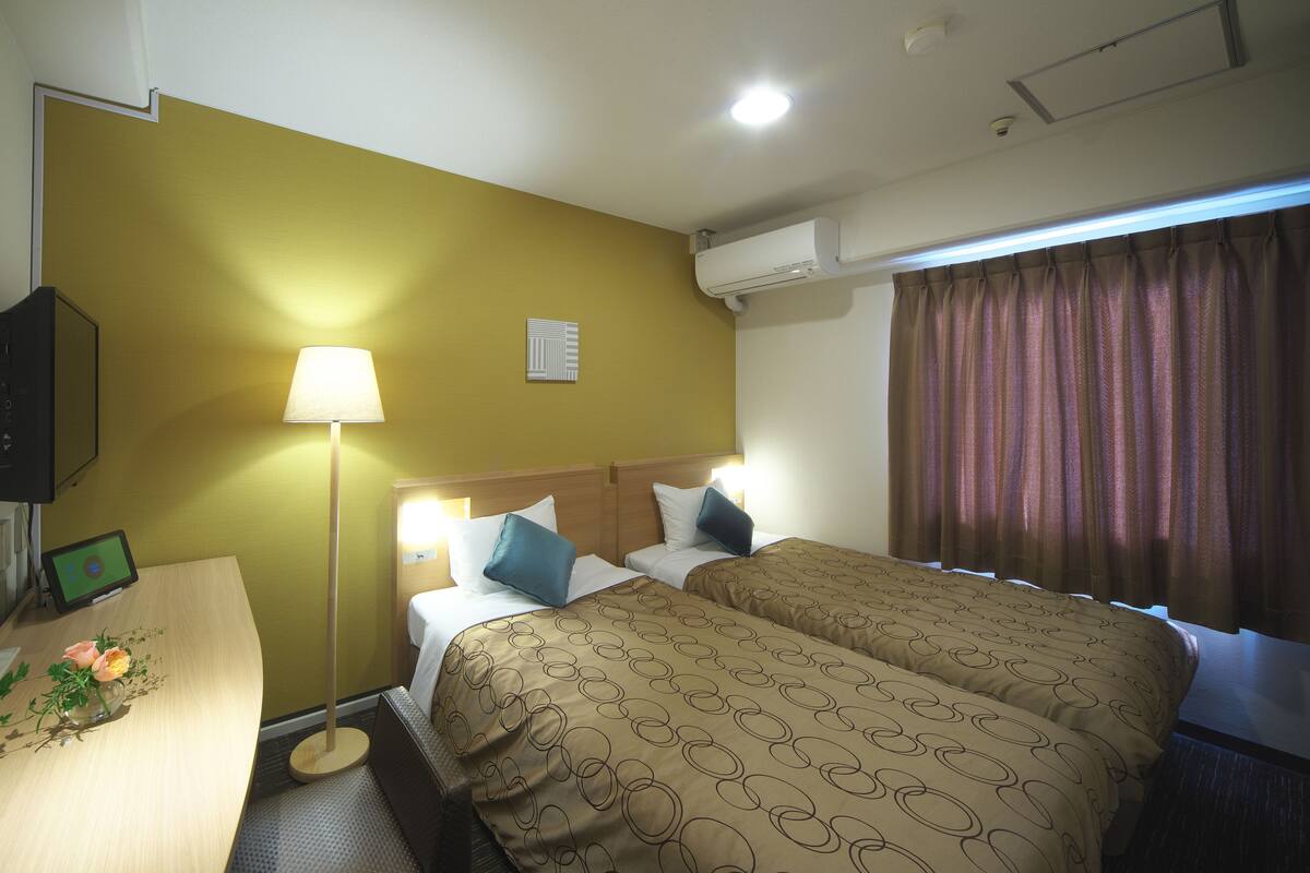 Ein Hotelzimmer mit zwei Betten, einem Schreibtisch mit Lampe, einem Fernseher und einem Fenster mit Vorhängen.