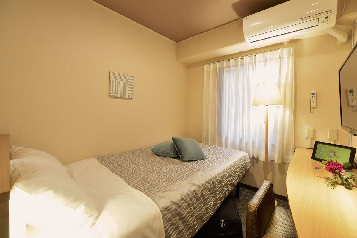 Ein Hotelzimmer mit einem Bett, einem Schreibtisch mit Computer, einer Klimaanlage und einem Fenster mit Vorhängen.