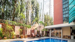 Une piscine dont le bord est recouvert de carreaux, entourée de plantes en pot et bordée d’un bâtiment à la façade rouge.