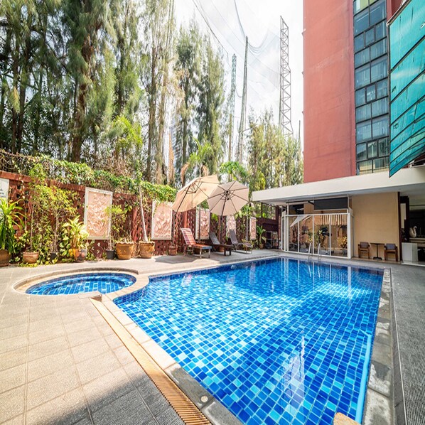 Una piscina con borde de azulejos, rodeada de plantas en macetas y un edificio de fachada roja.