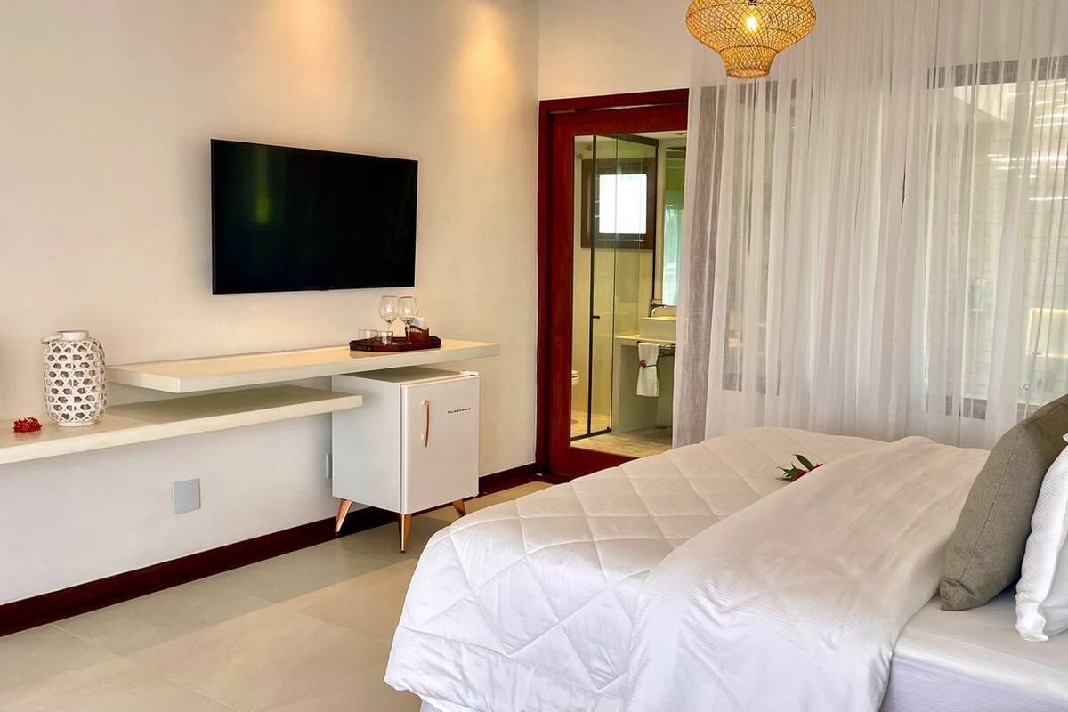 Ett modernt hotellrum med en stor säng, ett skrivbord med en platt-TV och ett badrum som syns genom en glasdörr.