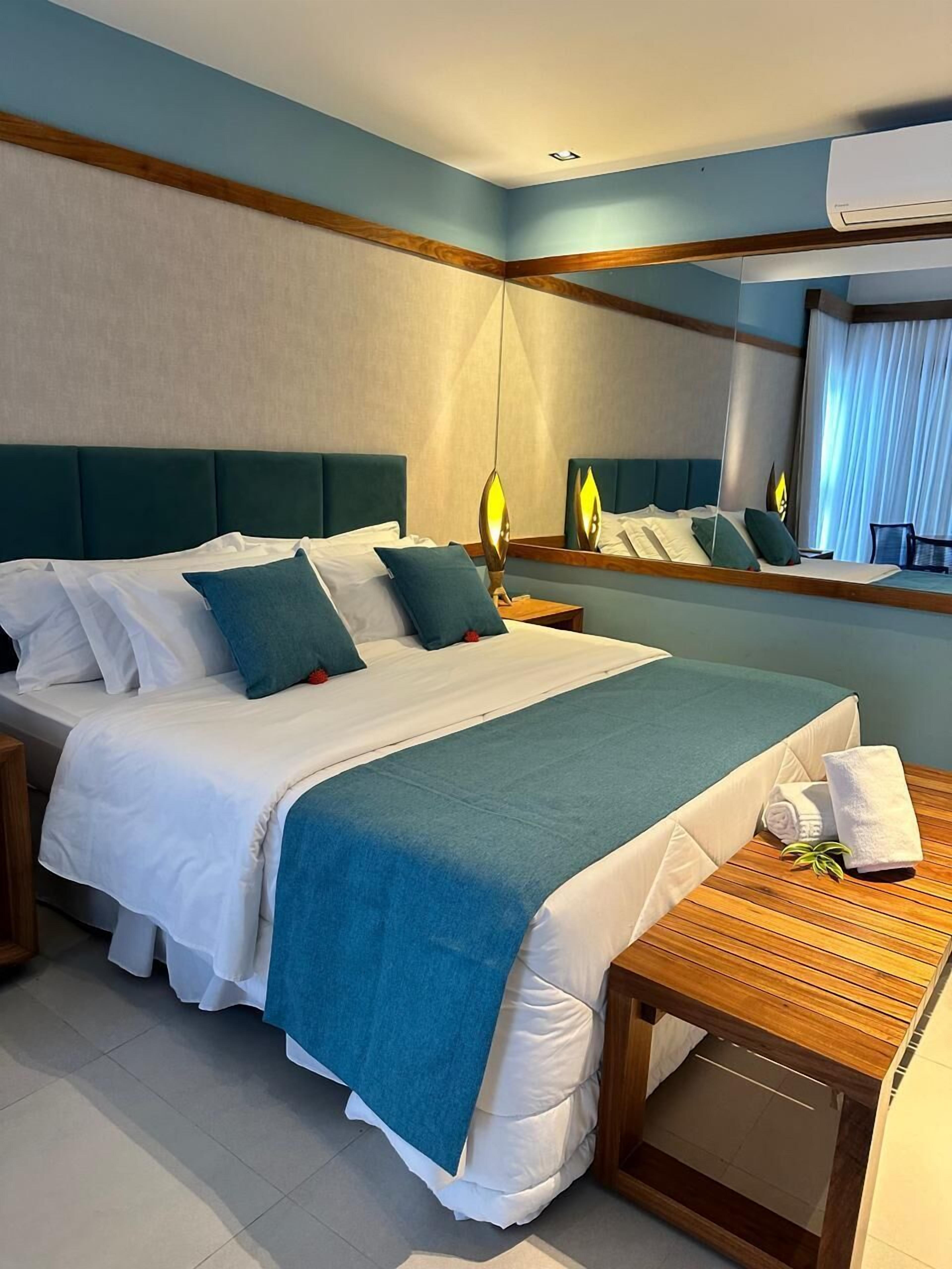 A hotel room with a large bed, a wooden nightstand, a bedside table with a towel, and a mirror reflecting the room.