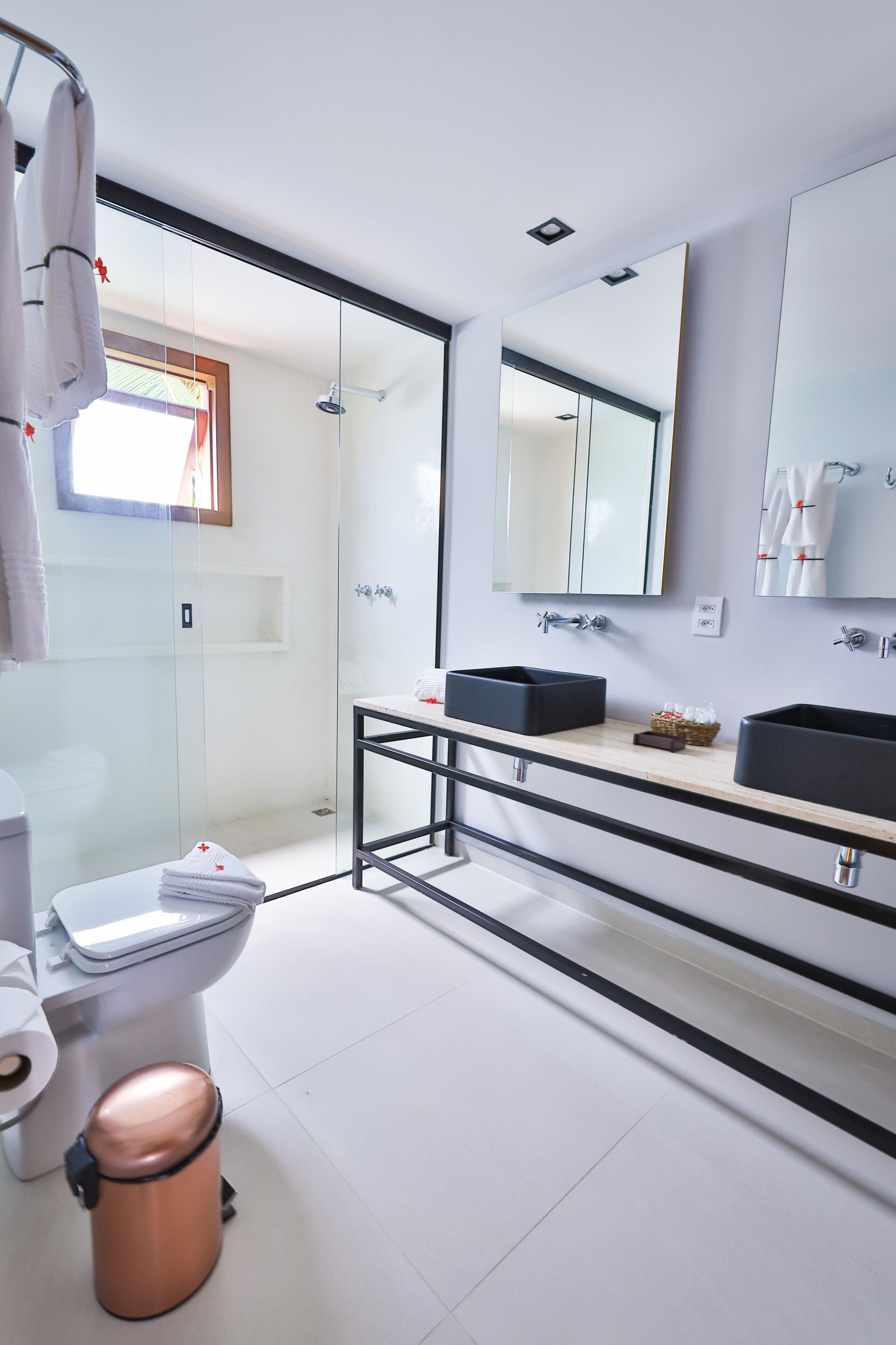 Un baño moderno con cabina de ducha de vidrio, inodoro blanco, cubo de basura de cobre y lavabo negro con un espejo arriba.