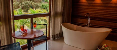Un baño moderno con una bañera independiente, una mesa redonda con bebidas y vistas a un follaje exuberante.