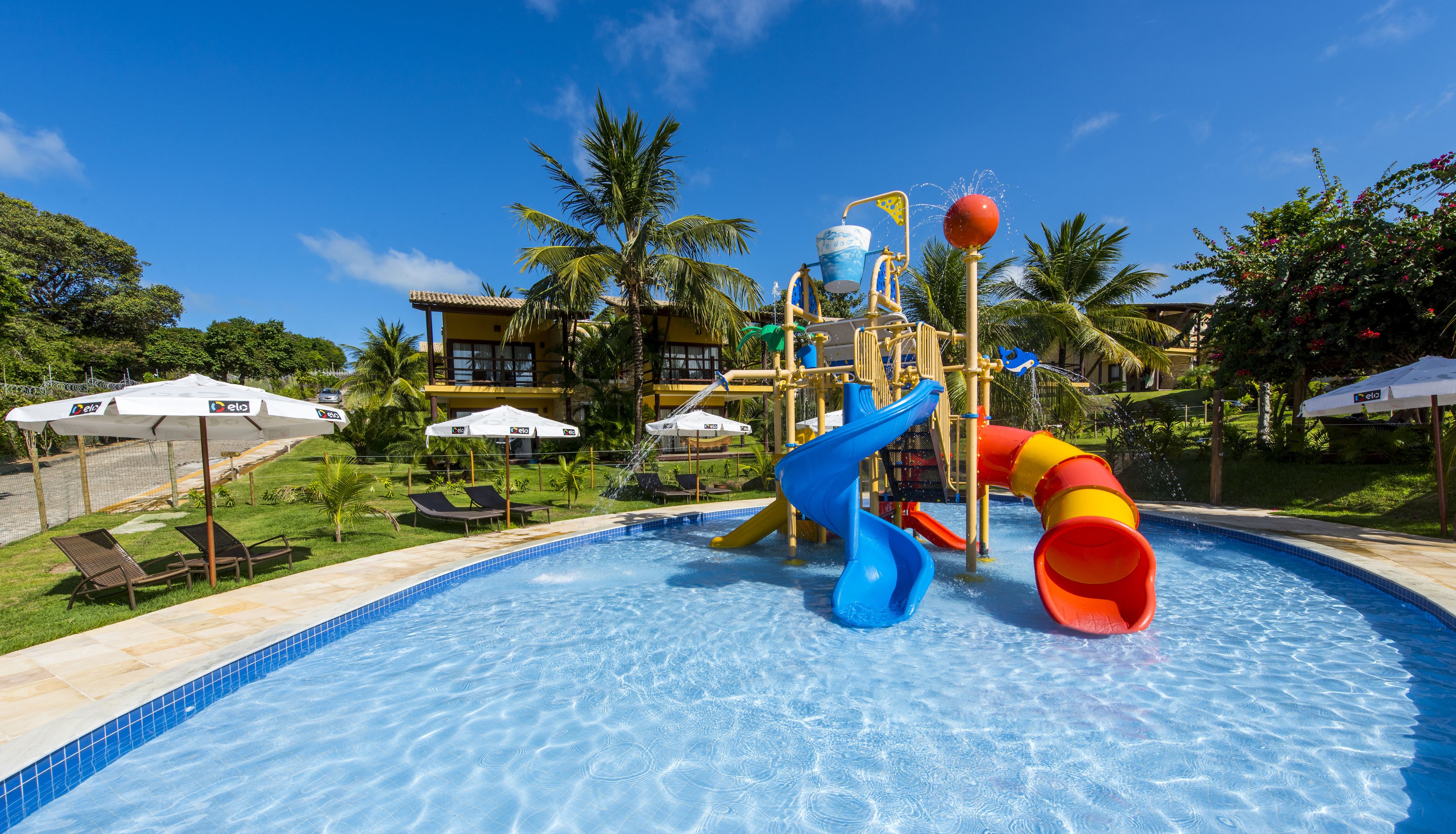 Una piscina con tobogán y estructura acuática. Hay sillones de descanso y sombrillas alrededor de la piscina.