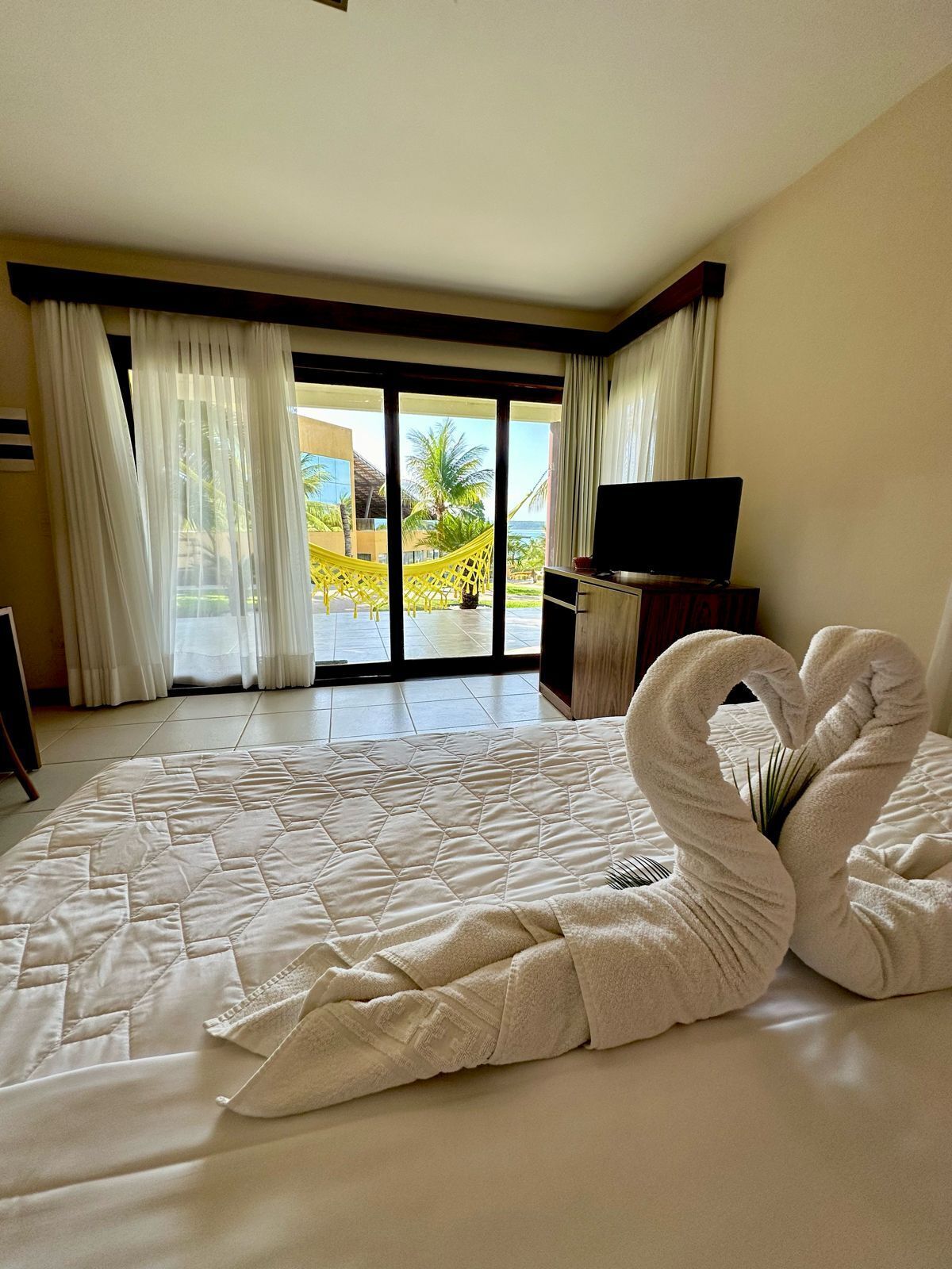 A hotel room with a large bed, a TV, and a view of palm trees and a beach.