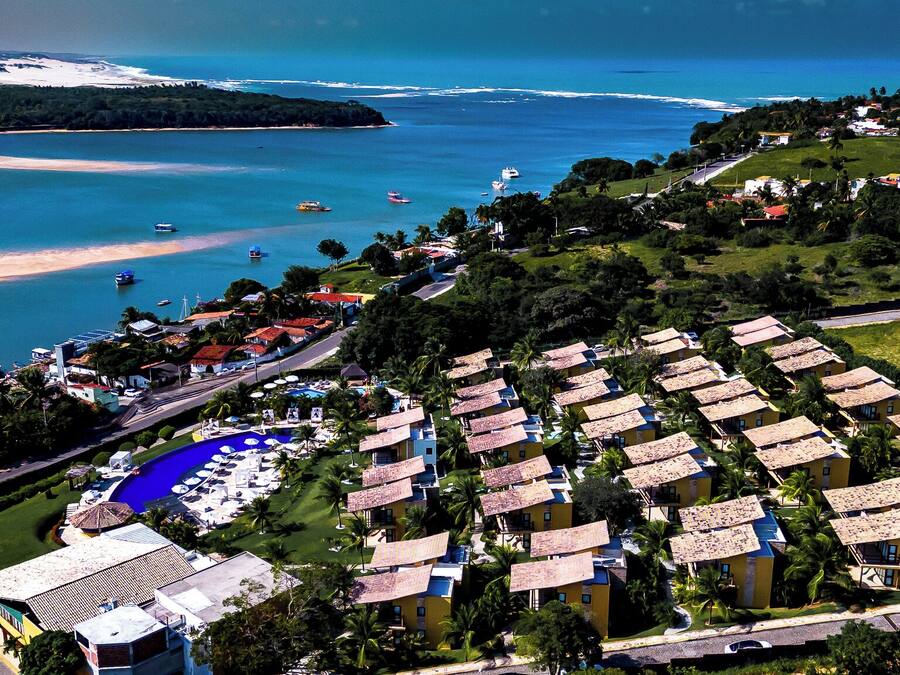 Un complejo turístico frente al mar con varias edificaciones tipo bungaló, un área de piscina central y una playa al fondo.