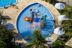 Una piscina con zona de juegos acuáticos, palmeras y sillas de descanso.