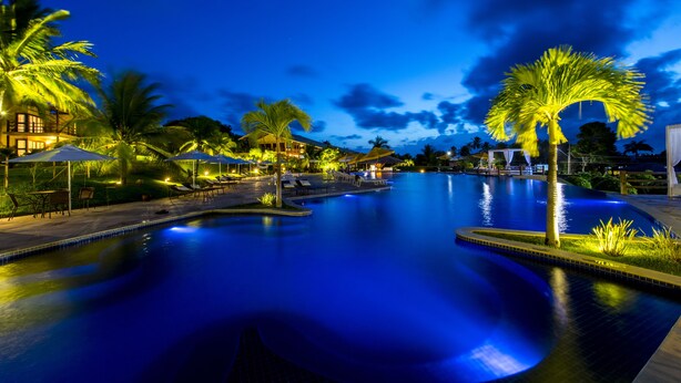 A nighttime swimming pool with palm trees and lounge chairs, surrounded by well-lit buildings. - Pipa Lagoa Hotel (Tibau do Sul)