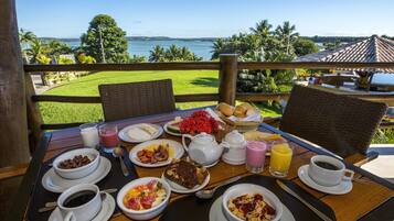 A breakfast table with various dishes, a view of the ocean, and a tropical landscape.