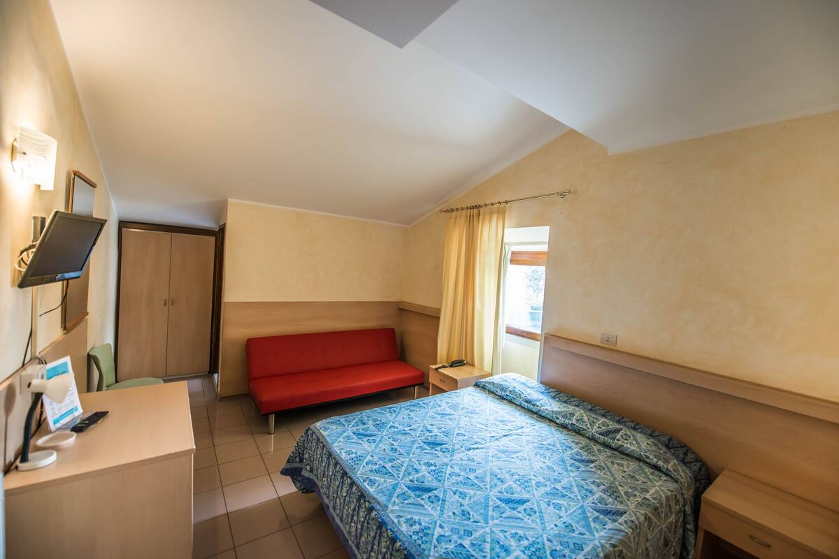 Une chambre d’hôtel avec un lit, un bureau, un canapé rouge et une fenêtre avec des rideaux.