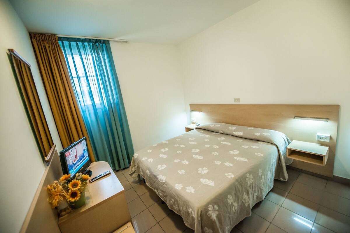 Une chambre d’hôtel avec un lit, des tables de chevet, un bureau avec un ordinateur et des rideaux.