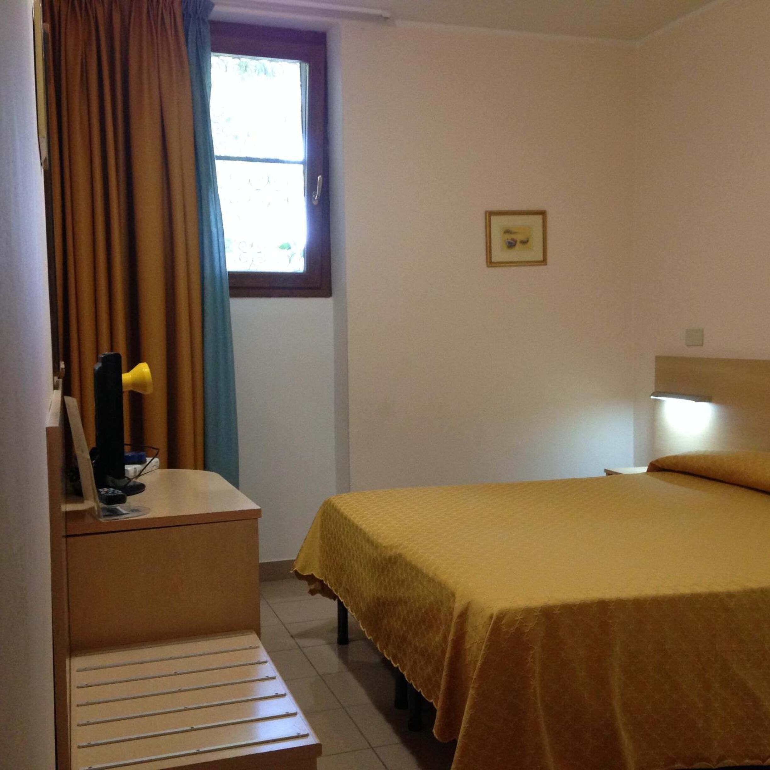 A small, simple room with a bed, a desk, a chair, and a window with curtains.