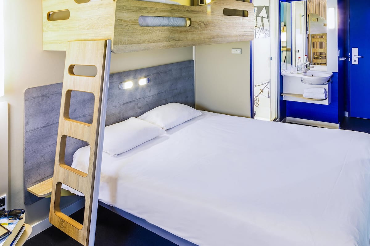 Una cama individual superpuesta con escalera de madera, cabecera gris y colchón blanco.