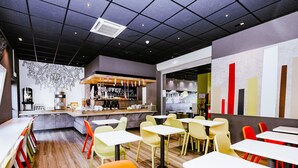 A modern cafe with a bar area, multiple seating options, and a decorative wall. - ibis budget Luxembourg Aéroport (Niederanven)