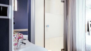 A hotel bathroom with a white sink, a glass shower enclosure, and a dark blue wall. - ibis budget Luxembourg Aéroport (Niederanven)