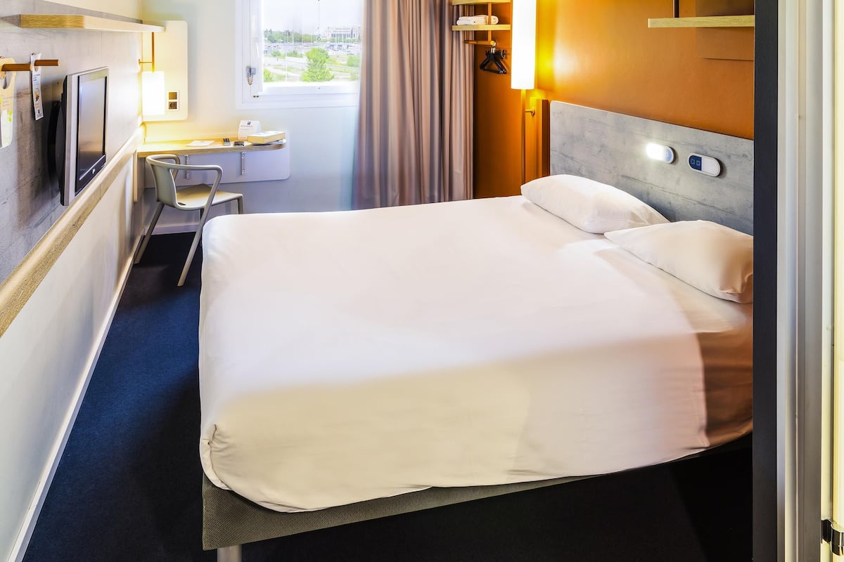 A hotel room with a bed, a desk with a chair, a television, and a window with curtains.
