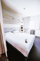 A modern hotel room with a large bed, a bedside table, a desk, and a chair.