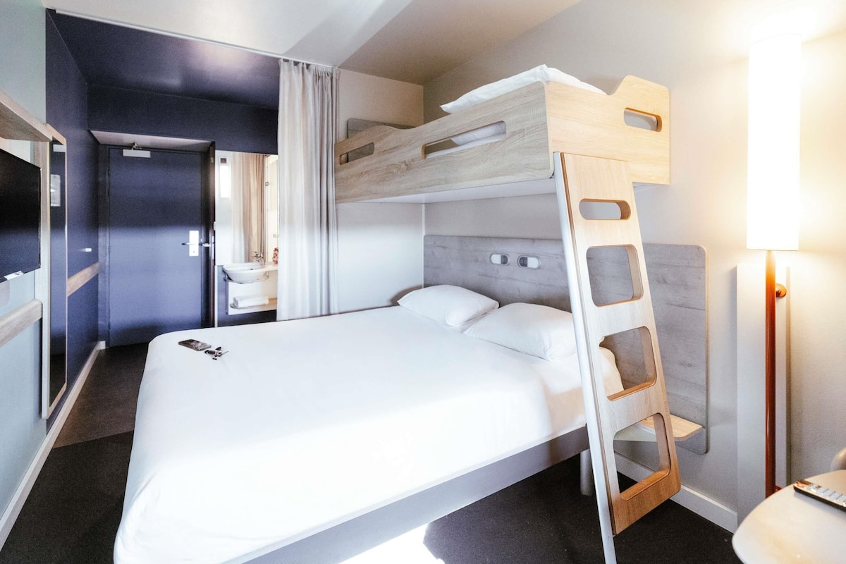 A bunk bed with a wooden headboard, a bedside table, a lamp, and a bathroom with a sink and mirror.