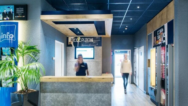 A reception area with a reception desk, a digital screen, and a sign that reads "RECEPTION." - ibis budget Luxembourg Aéroport (Niederanven)