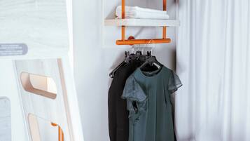 Una habitación ordenada con una cama, una mesa con un control remoto, una silla, un estante con toallas dobladas y un perchero con ropa.