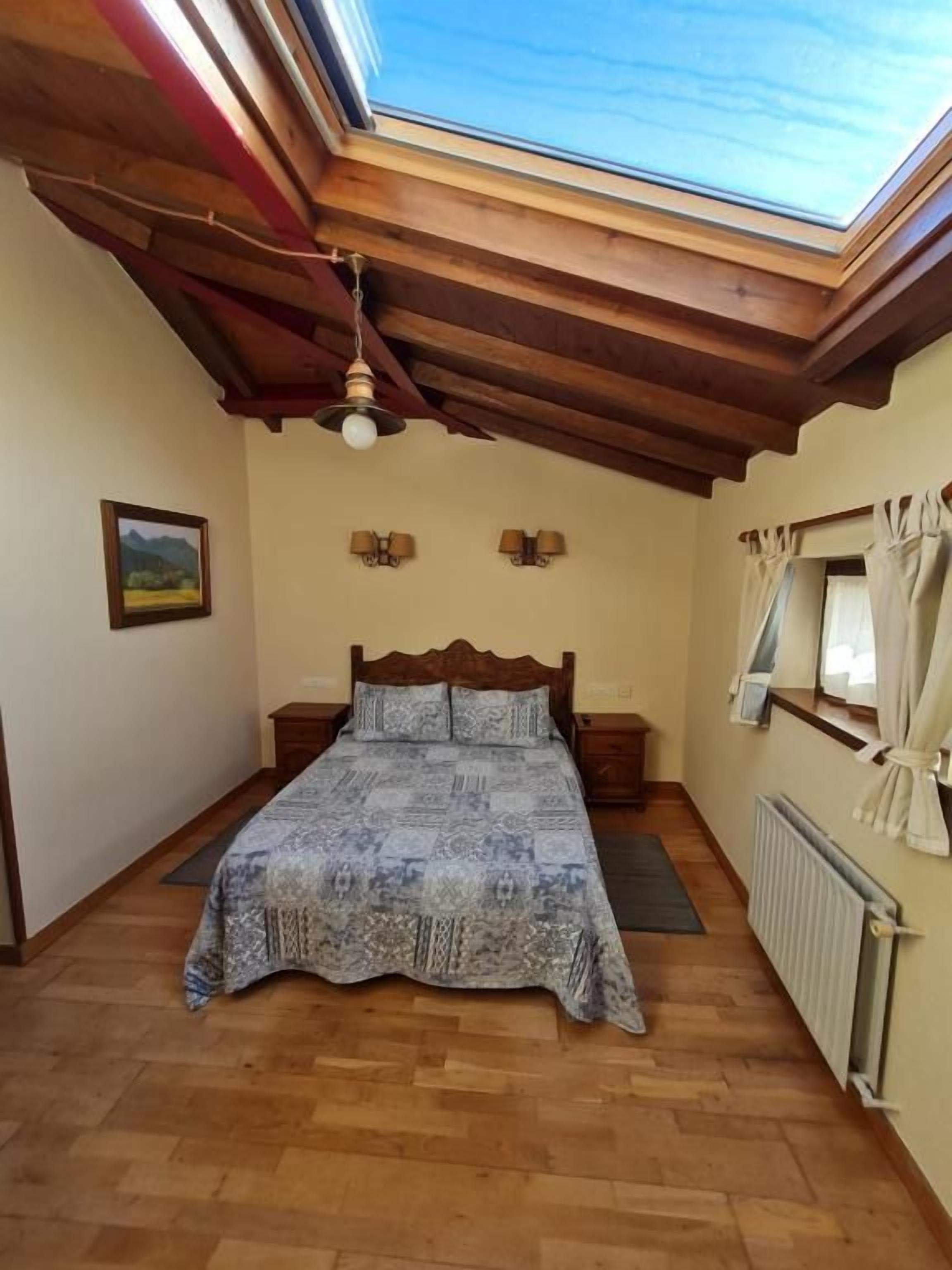 Un dormitorio con tragaluz, vigas de madera en el techo, una cama, mesitas de noche, lámparas de pared, un radiador y una ventana con cortinas.
