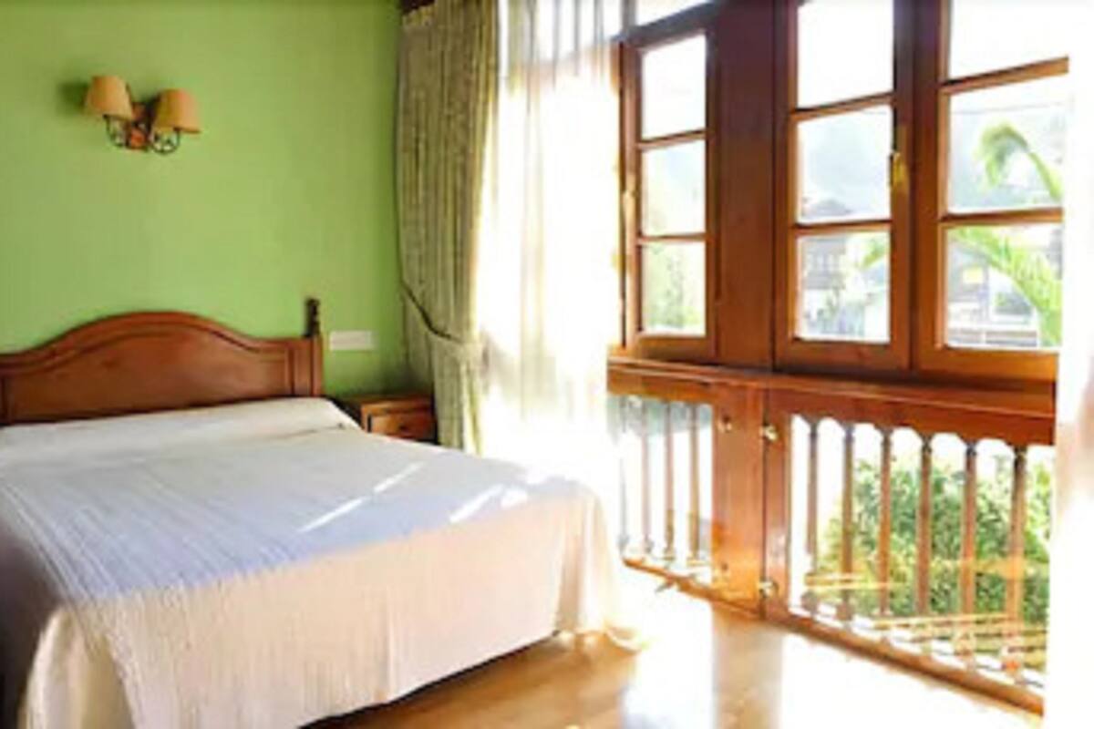 A bedroom with a wooden bed, a nightstand, a window with a view of greenery, and a balcony with a railing.