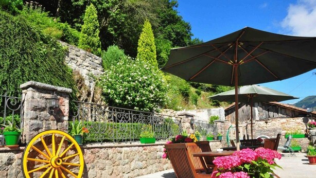 An outdoor seating area with a stone wall, a decorative yellow wheel, and a large umbrella providing shade. - La Balsa (Cangas de Onis)