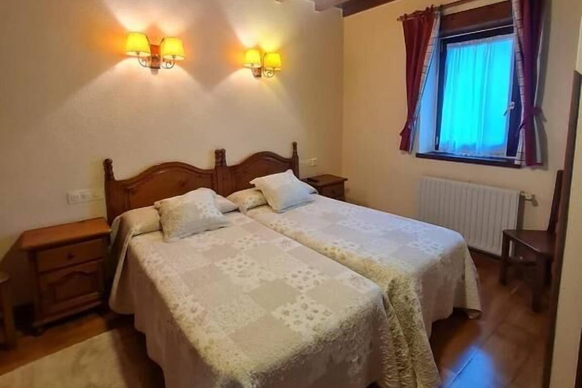 A bedroom with a wooden ceiling, two bedside lamps, a bed with a white bedspread, a window with curtains, and a radiator.