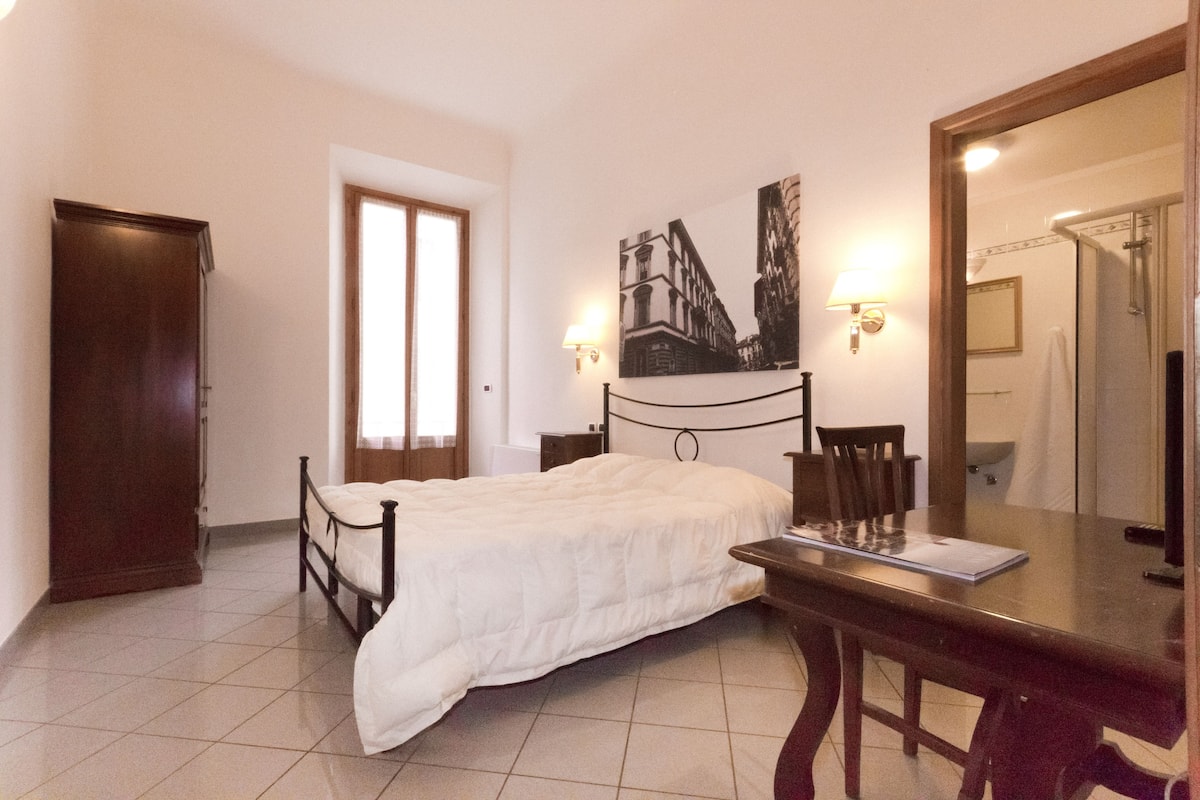 A bedroom with a bed, a desk, a chair, a wardrobe, a door, a wall-mounted lamp, and a mirror.