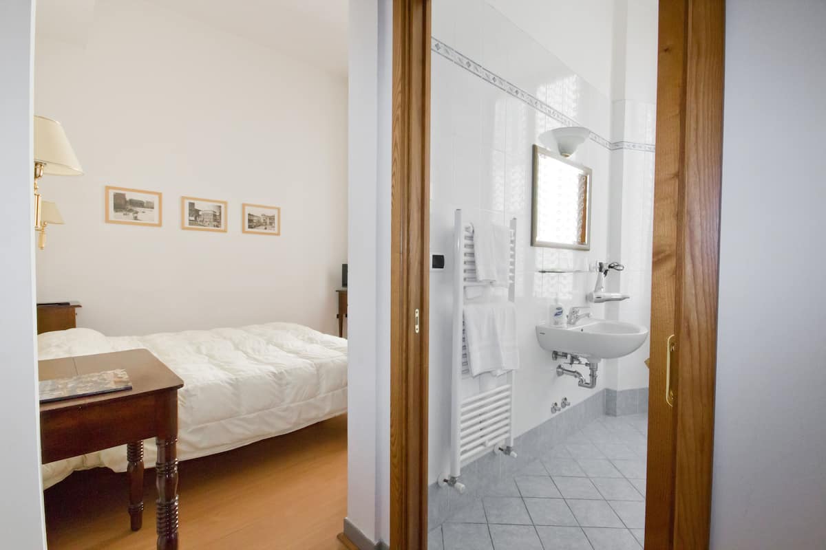 A bedroom with a bed, bedside table, and a nightstand. There is a bathroom with a sink, mirror, and towel rack.
