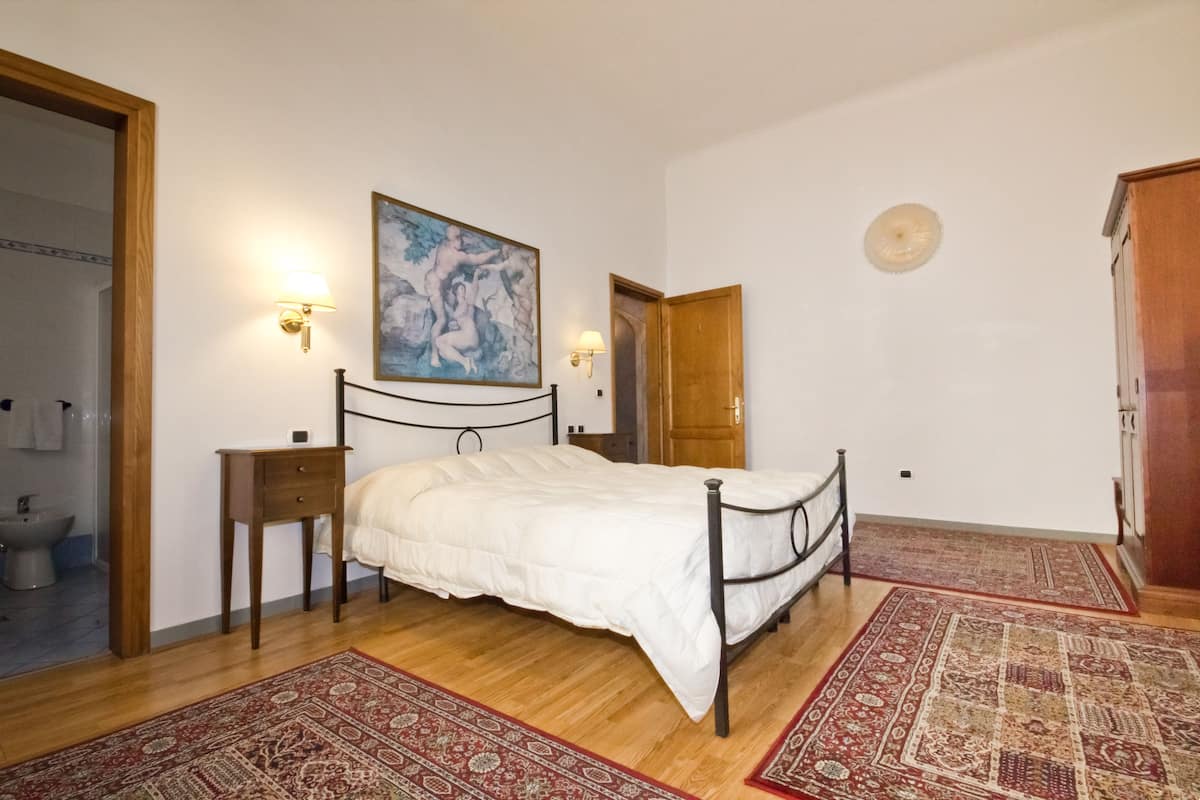 A bedroom with a bed, two bedside tables, a nightstand, a painting, a door, and a window.