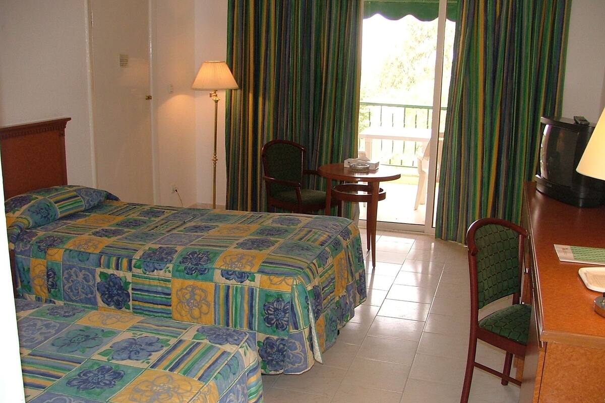 Habitación de hotel con dos camas, un escritorio, una silla, una lámpara y vistas al exterior.