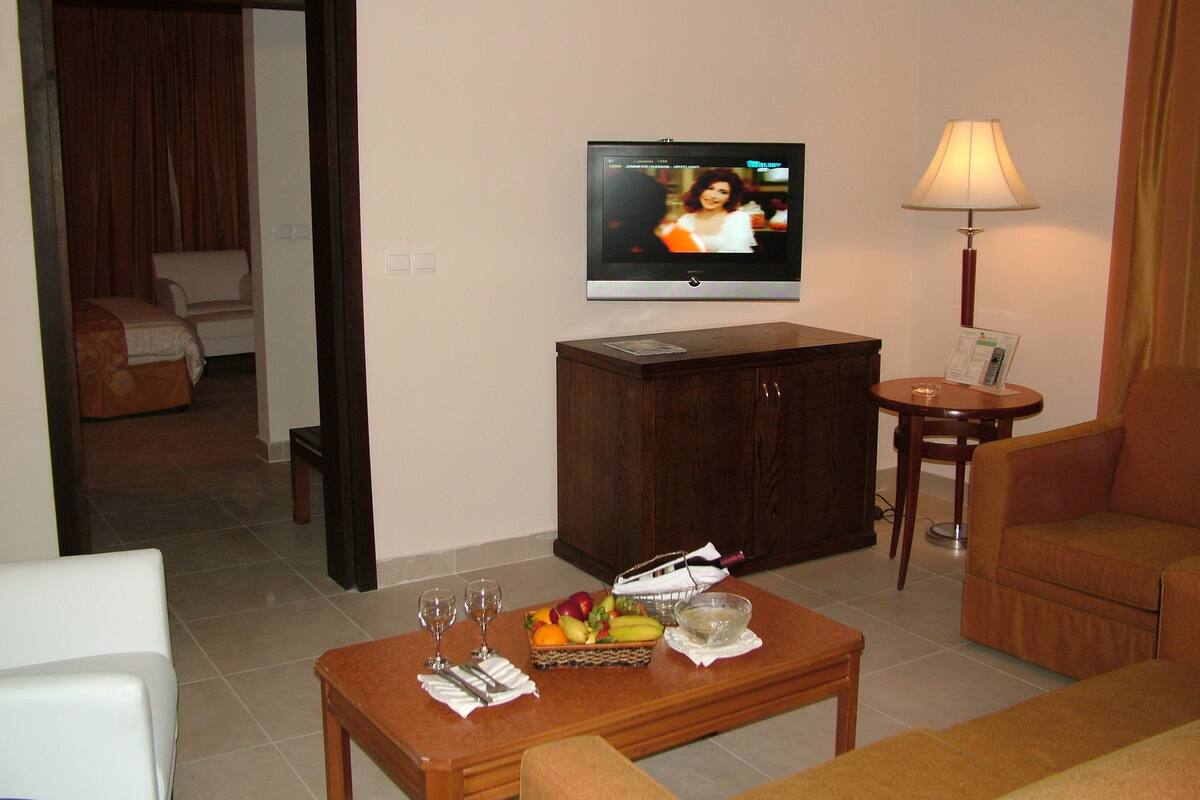 Habitación de hotel con televisor, una mesa de comedor con fruta y copas de vino, una lámpara y un sofá.