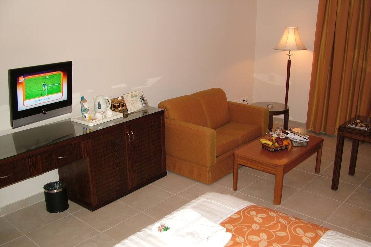 Habitación de hotel con televisor de pantalla plana, sofá, una mesita con fruta, lámpara y cama con manta estampada.
