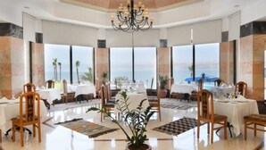A spacious dining area with a central chandelier, large windows offering a view of the sea, and a polished floor. - Dead Sea Spa Hotel (Sweimeh)