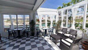 A covered outdoor seating area with checkered flooring, tables, and chairs, overlooking a body of water and buildings. - Cala Bona & Mar Blava Hotels (Ciutadella de Menorca)