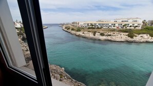A coastal view from a window overlooking a clear blue sea, a rocky shoreline, and a row of buildings with balconies. - Cala Bona & Mar Blava Hotels (Ciutadella de Menorca)