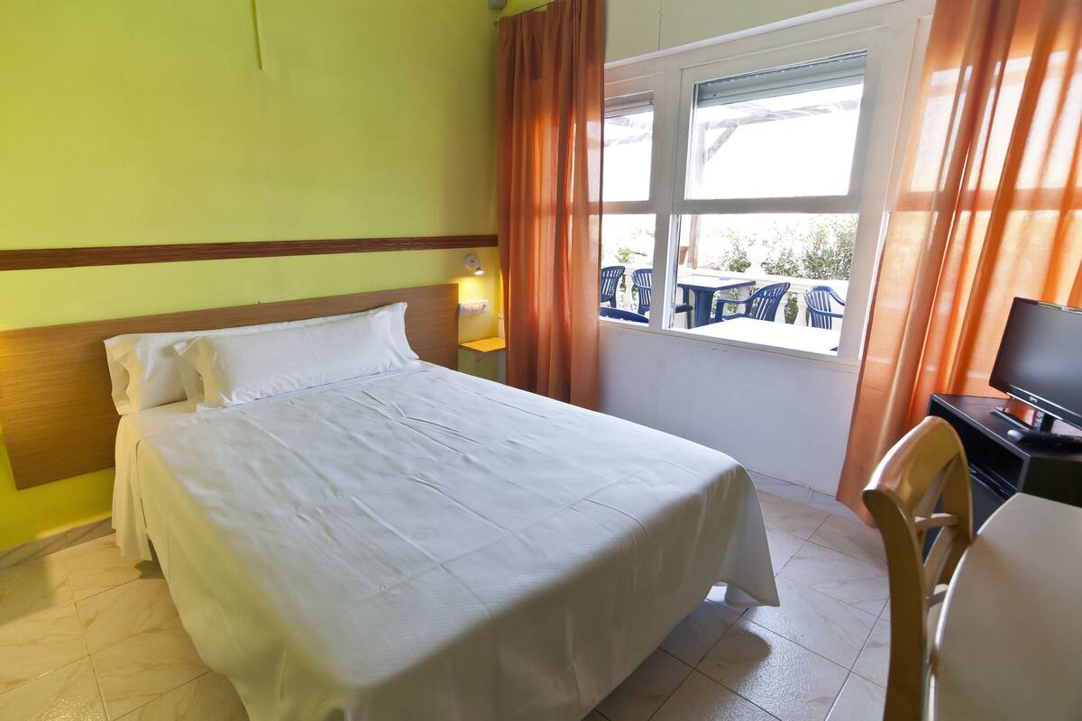 Ein Hotelzimmer mit Bett, Schreibtisch mit Computer, Fenster mit Vorhängen und Blick nach draußen.