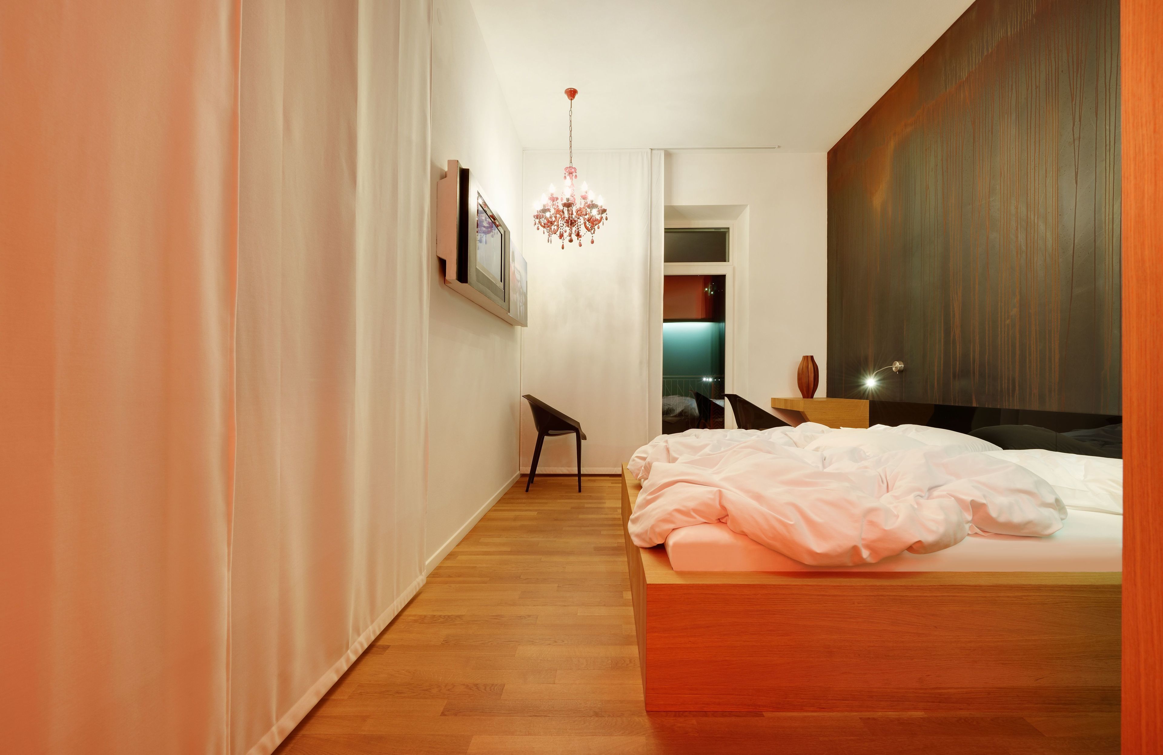 A modern bedroom with a wooden bed, a hanging chandelier, and a view into another room.