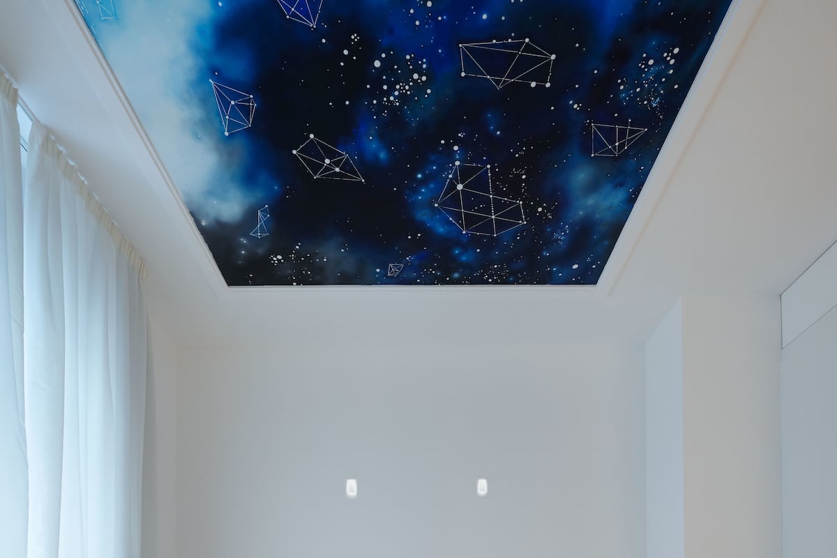 Un dormitorio con techo que simula un cielo estrellado, cama con ropa de cama blanca y dos luces fijadas a la pared.